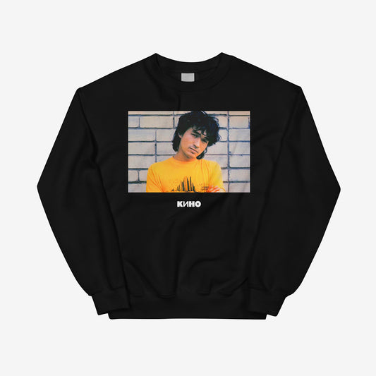 Rare archival photo of Viktor Tsoi in a yellow T-shirt against a white brick wall with a little KINO logo underneath, printed on the front of a heavyweight black unisex sweatshirt with a KINO logo on the back neck side, from GruppaKino.com. Front side.