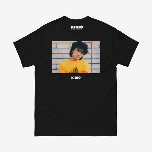 Rare archival photo of Viktor Tsoi in a yellow T-shirt against a white brick wall, printed on the front of a heavyweight black unisex t-shirt with a KINO logo under the photo and on the inside neck label, from GruppaKino.com.